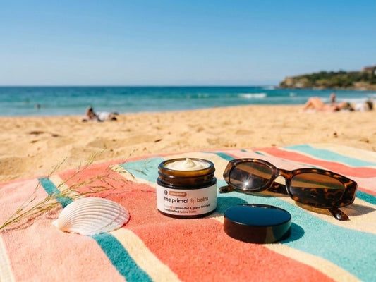 Open jar of The Primal unscented whipped tallow balm showing the creamy balm, on a striped beach towel with sunglasses at a sunny Sydney beach